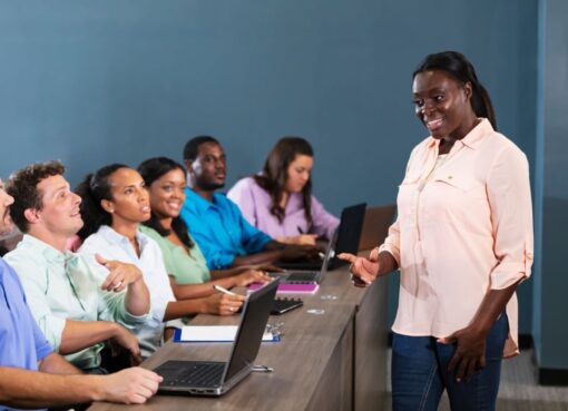 A mature African-American woman teaching a talent acquisition training seminar. The professionals are sitting in a row at a long table, with their note pads and laptop computers, looking up at the instructor standing in front of them.